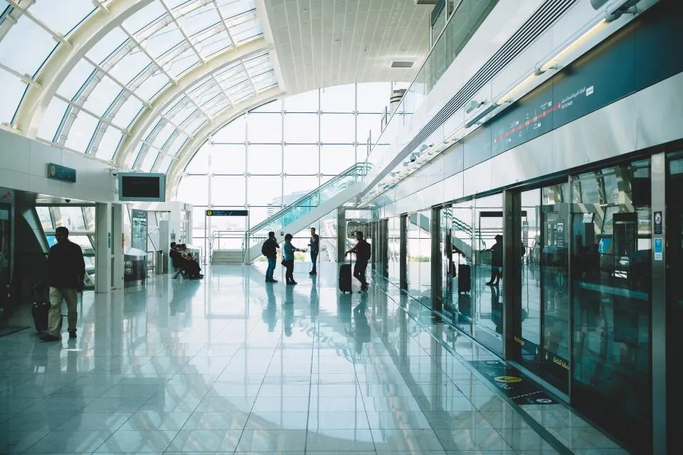 Passengers wheeling suitcases through a bright glass-roofed airport terminal