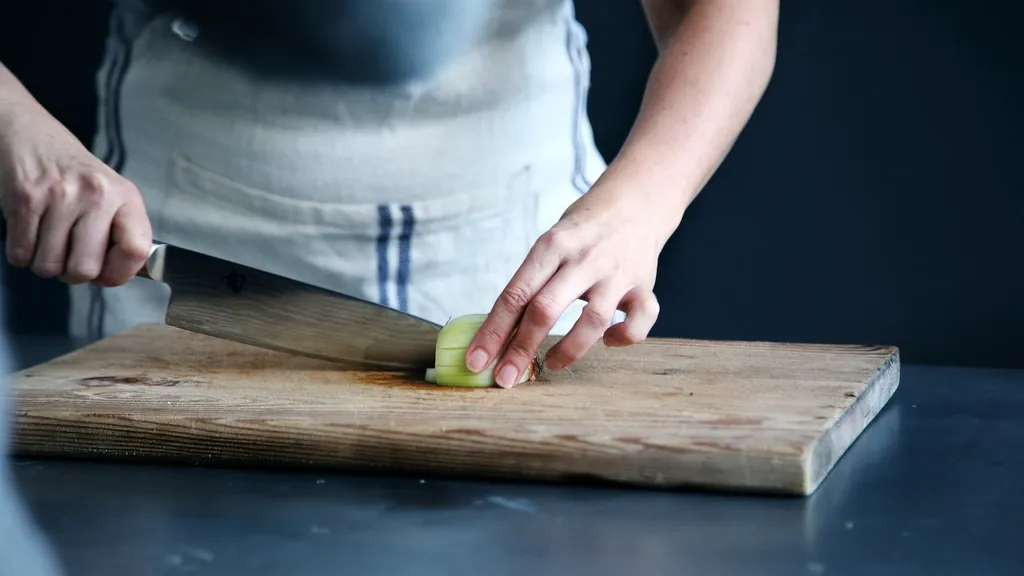 A cook slicing an onion with a chef’s knife on a wooden board, fingertips curled back