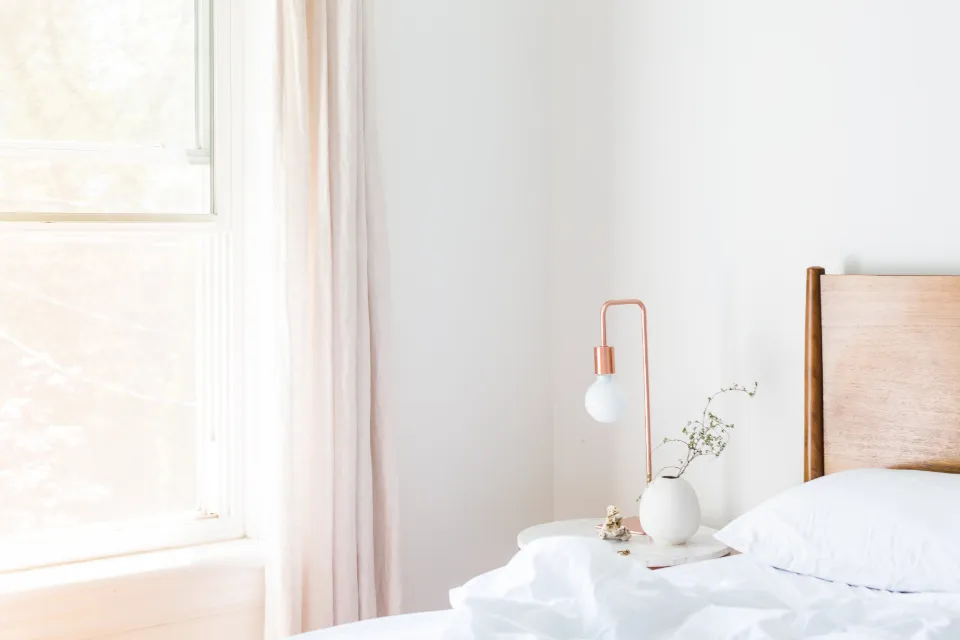 A sunlit bedroom with white bedding, a pale linen curtain at the window and a copper lamp on the bedside table