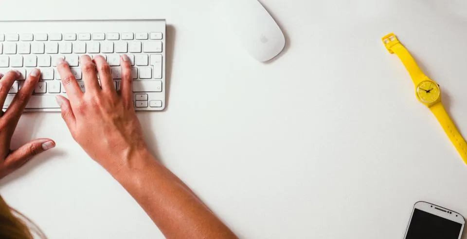 Hands typing on a white keyboard beside a mouse, a watch and a phone on a plain desk