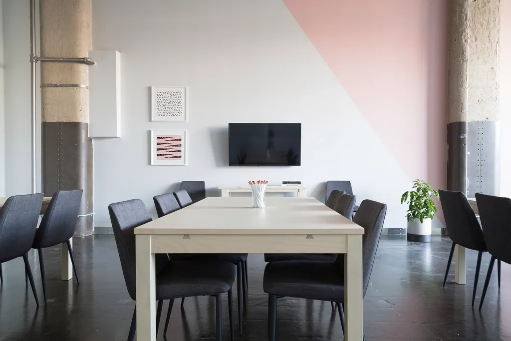 An empty meeting room with a long pale table, dark chairs and a wall-mounted screen