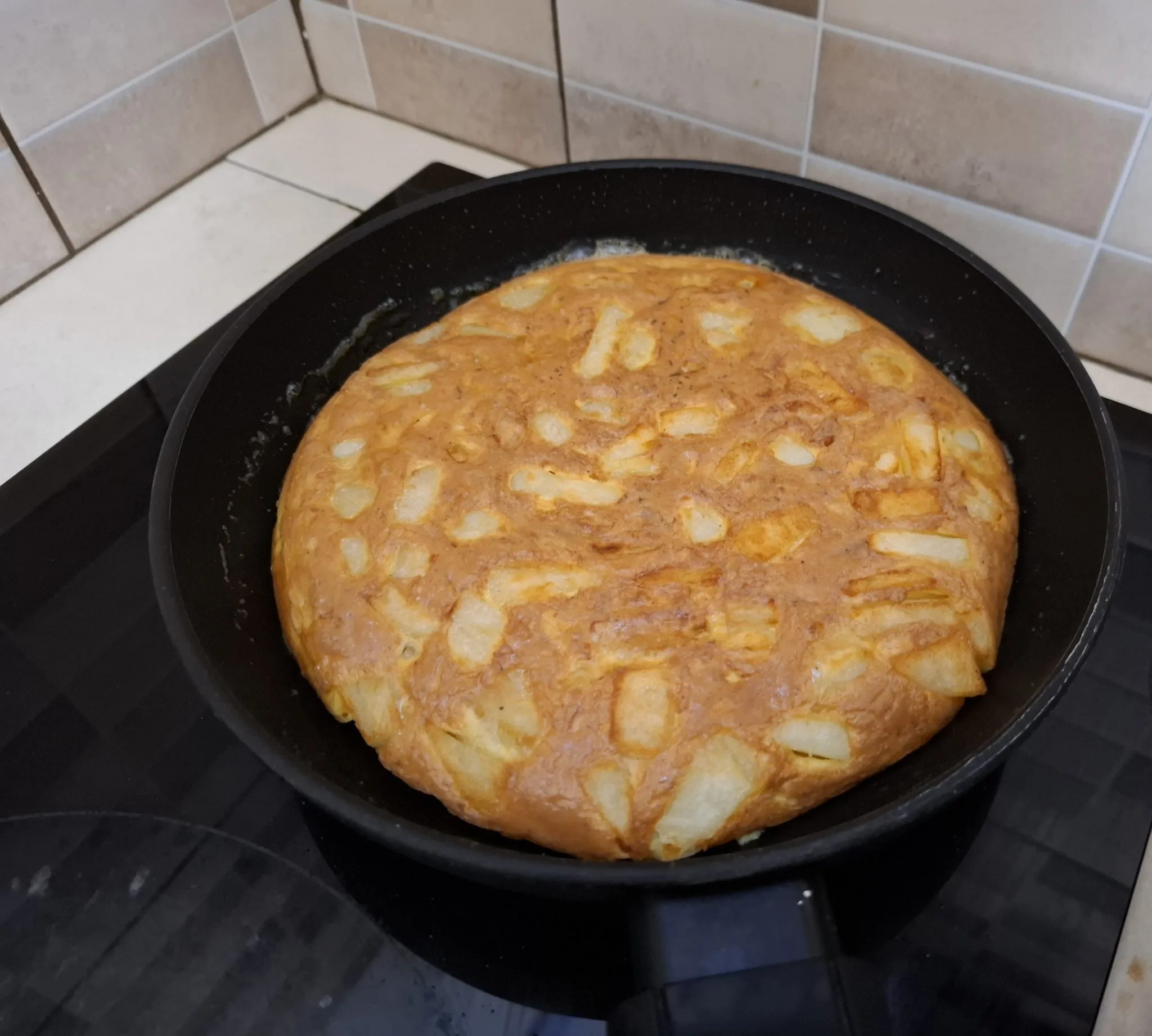 A thick potato omelette cooking in a black frying pan on a hob