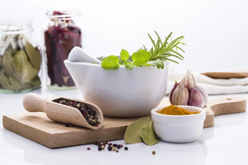 A white mortar and pestle holding fresh herbs, beside peppercorns, garlic and a dish of ground turmeric