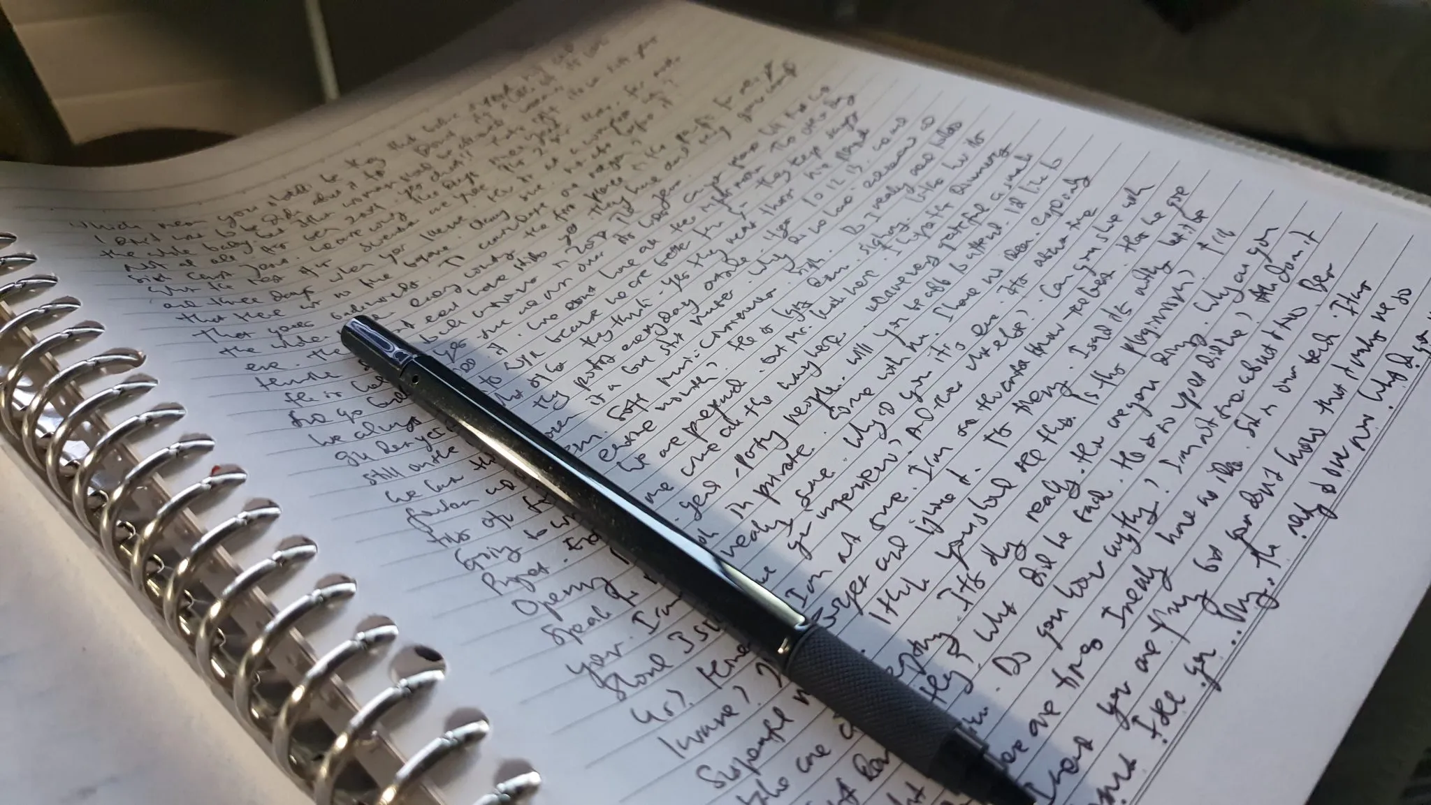 A spiral-bound notebook filled with handwriting, a pen resting across the page