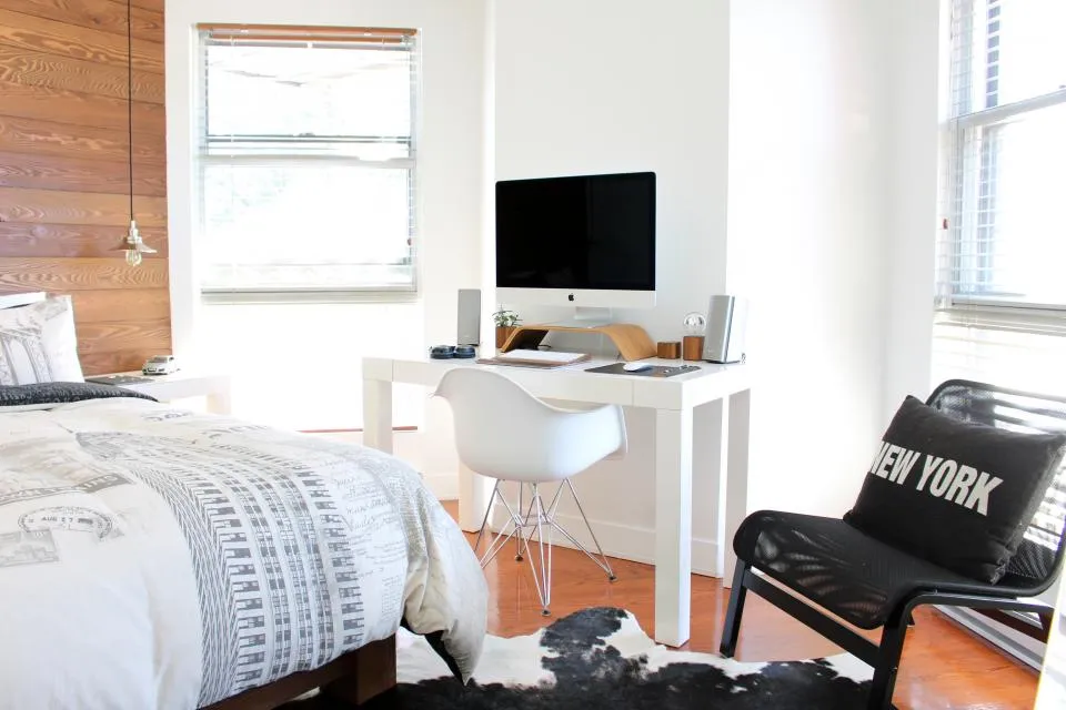 A desk with a monitor and chair set up in the corner of a bedroom beside a window
